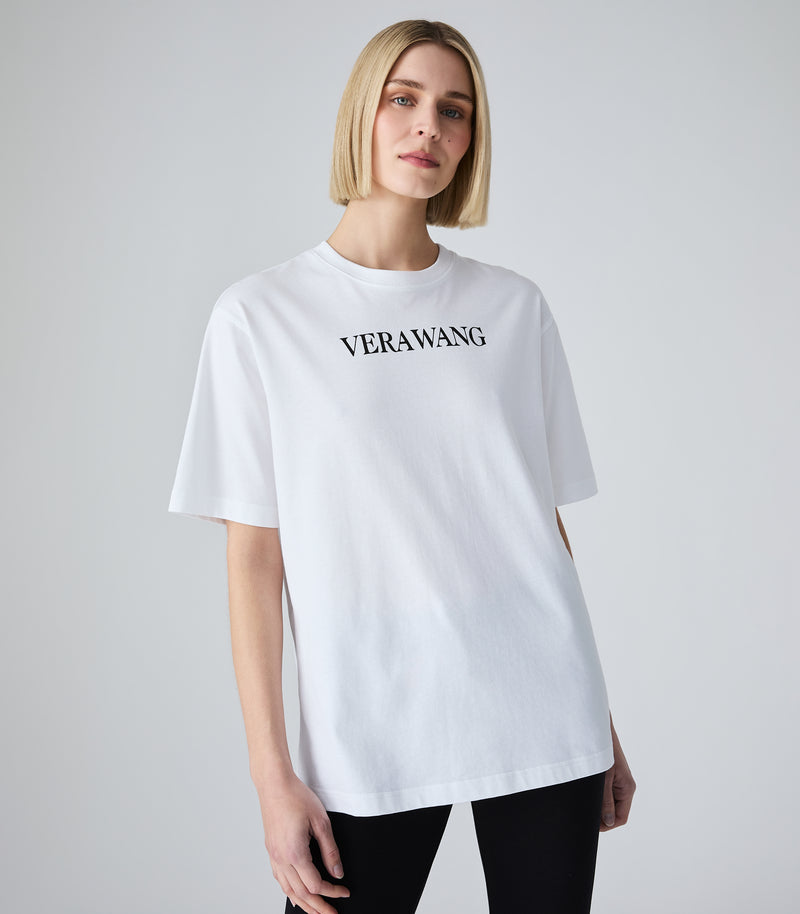 Three-quarter length view of a model wearing an oversized, loose-fit white t-shirt featuring the classic 'VERA WANG' brand logo printed in black across the upper chest, designed with wide short sleeves and a crew neckline, paired with black pants against a light grey background.