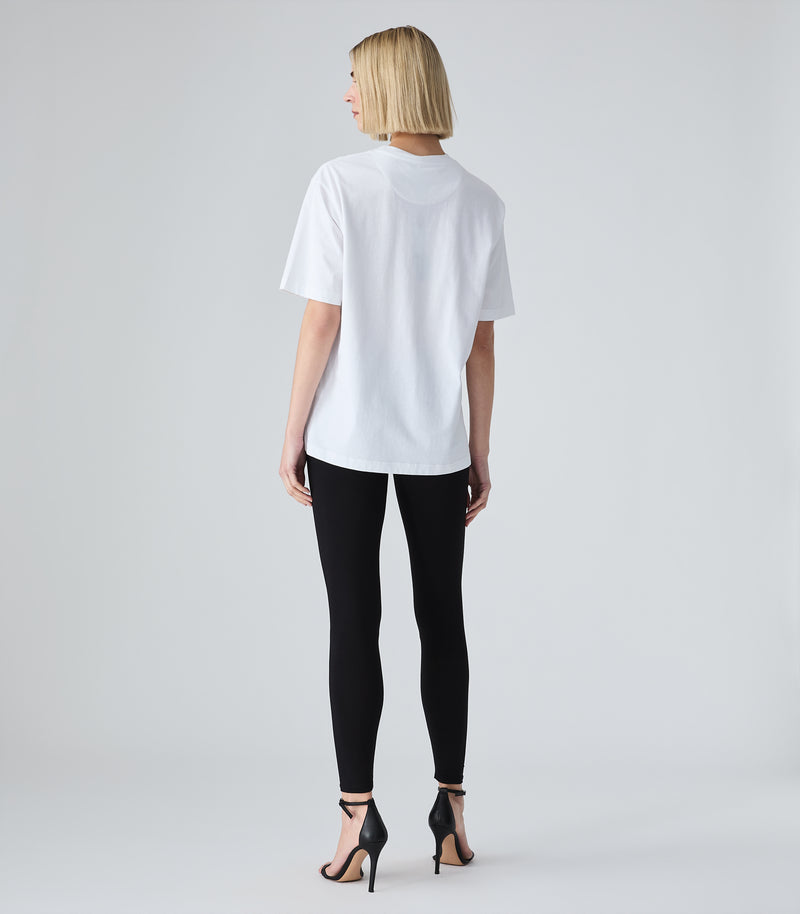 Full-length back view of a model wearing an oversized white cotton t-shirt, showcasing a relaxed silhouette, wide short sleeves, and a subtle crescent-shaped yoke seam below the crew neck collar. The look is styled with black fitted leggings and black ankle-strap stiletto sandals against a light grey background.
