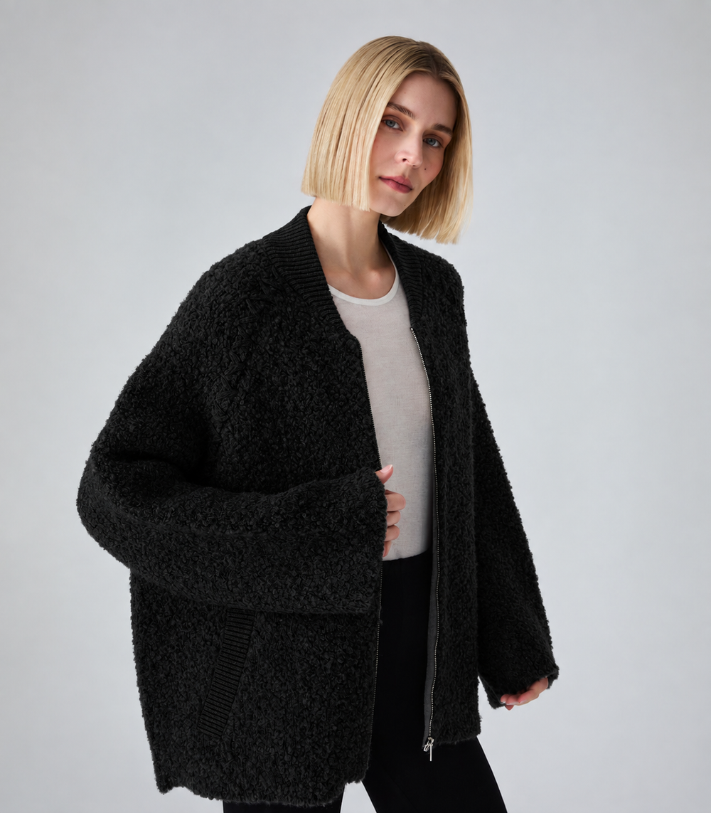 A woman with a short blonde bob wearing a black textured teddy fleece zip-up cardigan over a light gray top and black pants, photographed against a neutral light gray studio background.
