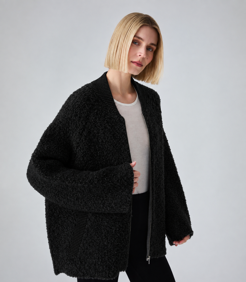 A woman with a short blonde bob wearing a black textured teddy fleece zip-up cardigan over a light gray top and black pants, photographed against a neutral light gray studio background.