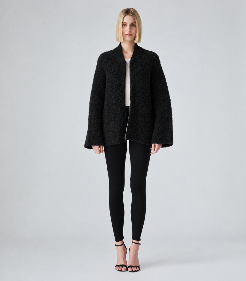 A woman stands against a light gray studio background wearing a black textured bouclé jacket over a light top, black leggings, and black heeled sandals.