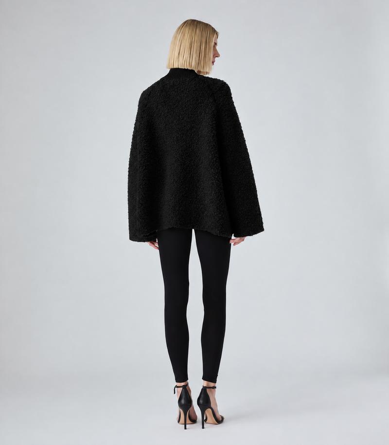 A woman viewed from the back wearing a black textured boucle-style jacket, black leggings, and black high-heeled sandals against a light gray studio background.
