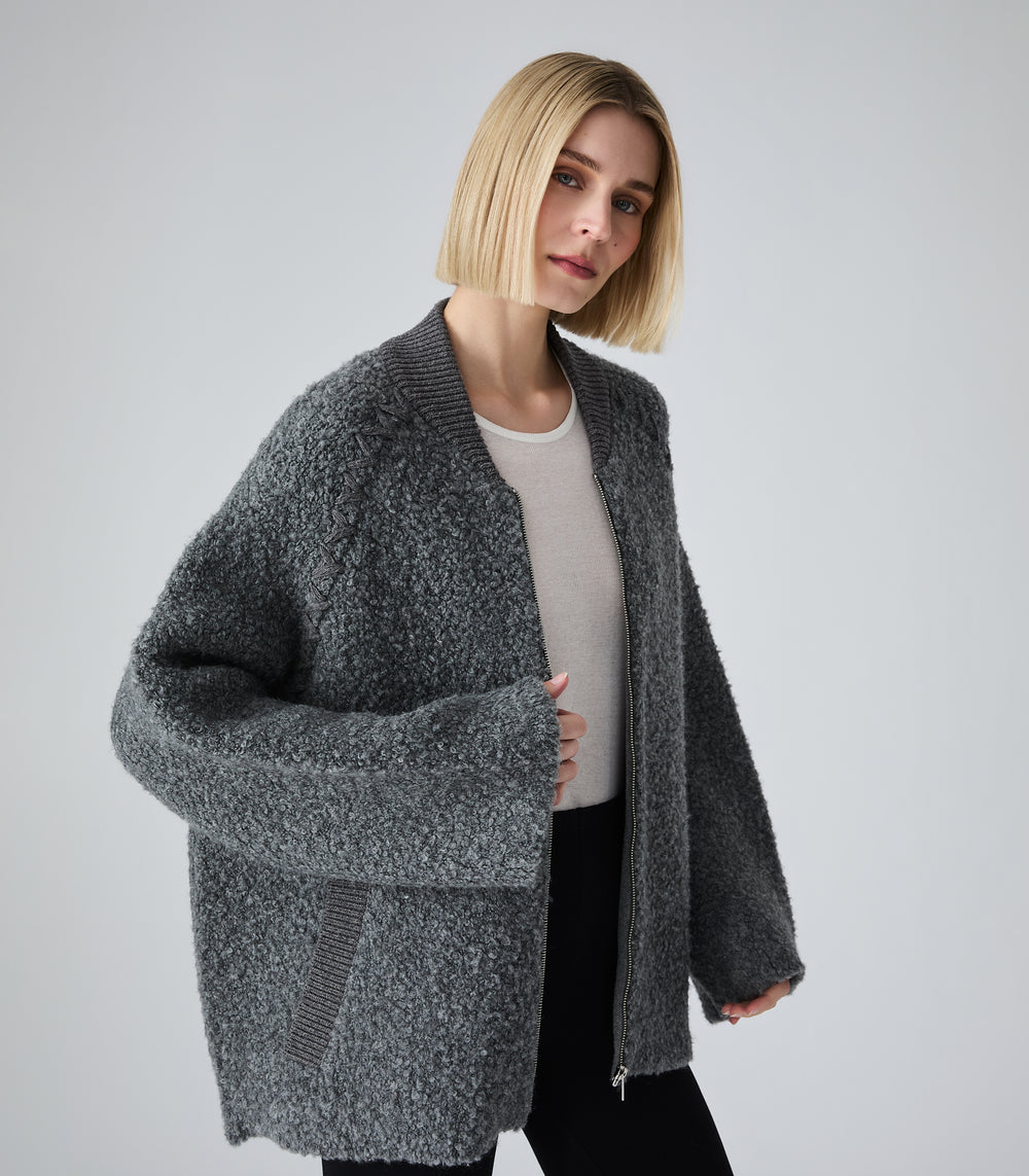 A woman with a short blonde bob wearing a dark gray marled zip-up cardigan over a light gray top and black pants, photographed against a plain light gray studio background.