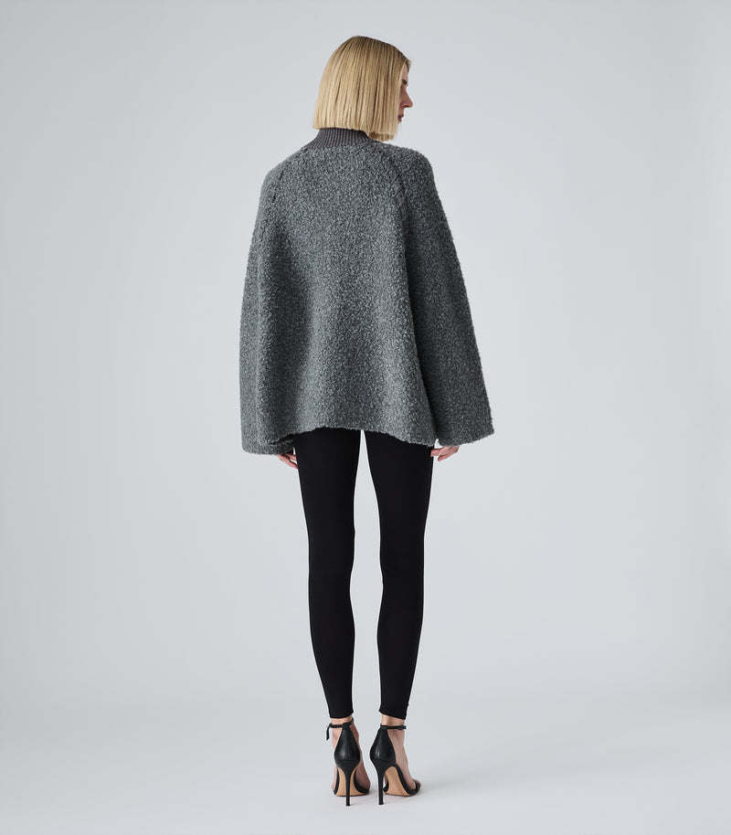 A woman shown from the back wearing a dark gray bouclé cape jacket, black leggings, and black high-heeled sandals against a light gray studio background.