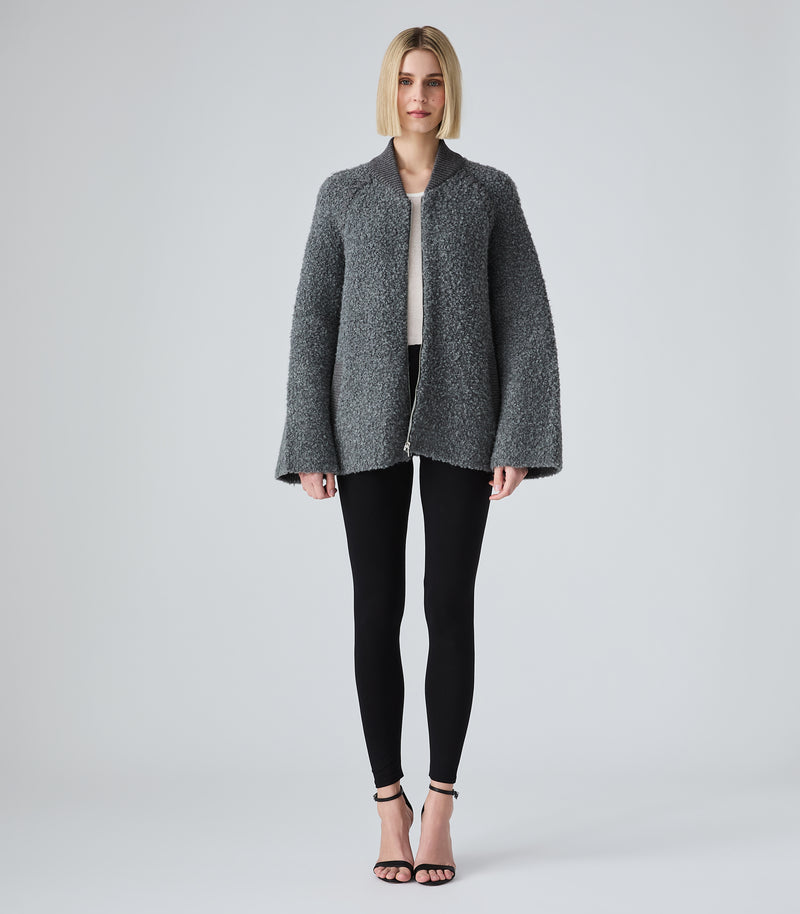 A woman stands against a light gray studio background wearing a textured charcoal gray open-front jacket with wide flared sleeves, black leggings, and black heeled sandals.