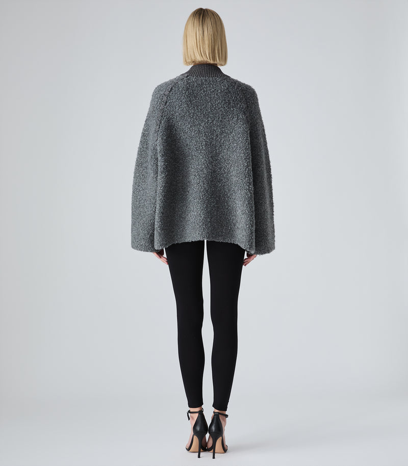 A woman viewed from the back wearing a dark gray textured boucle jacket, black leggings, and black high-heeled sandals against a light gray studio background.