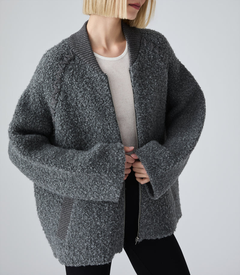A person wearing an oversized dark gray bouclé zip-front cardigan over a light beige top and black pants, photographed against a plain light gray studio background.