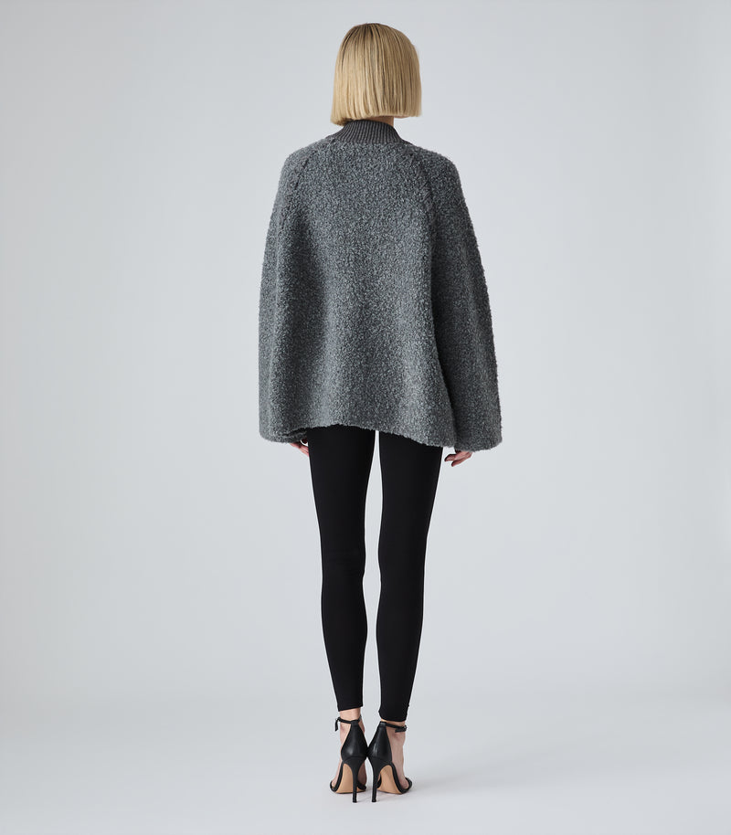 A model viewed from the back wearing a textured gray boucle-style cropped jacket, black leggings, and black high-heeled sandals against a light gray studio background.
