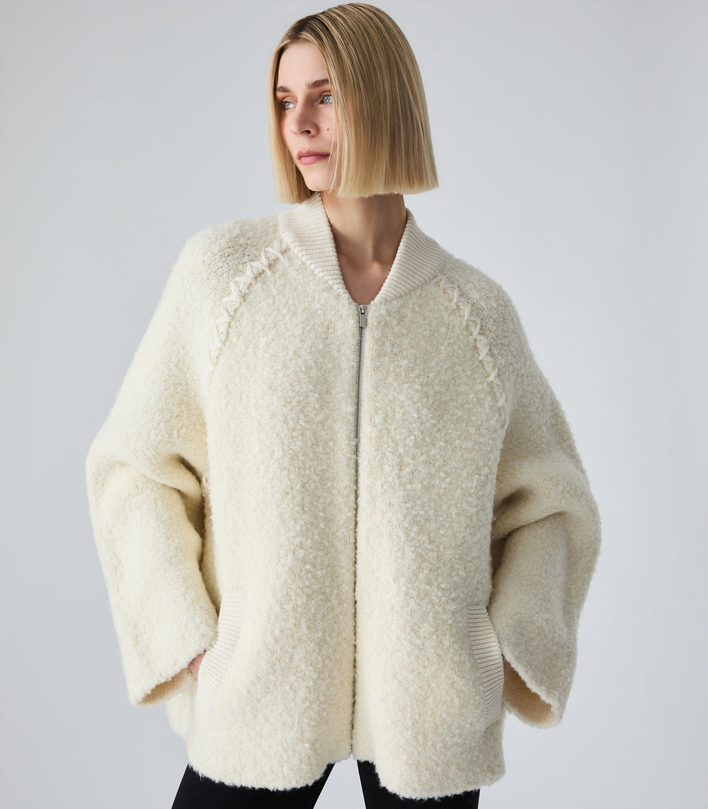 A woman with a short blonde bob wearing a cream-colored, textured teddy fleece zip-up jacket with ribbed collar and cuffs, paired with black pants, against a light gray studio background.