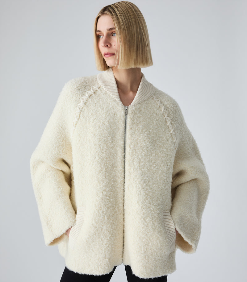 A woman with a short blonde bob wearing a cream-colored, textured teddy fleece zip-up jacket with ribbed collar and cuffs, paired with black pants, against a light gray studio background.