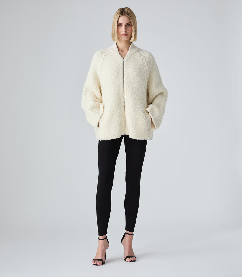 A woman modeling a cream-colored, textured teddy fleece zip-up jacket with a stand collar, paired with black leggings and black high-heeled sandals, against a light gray studio background.