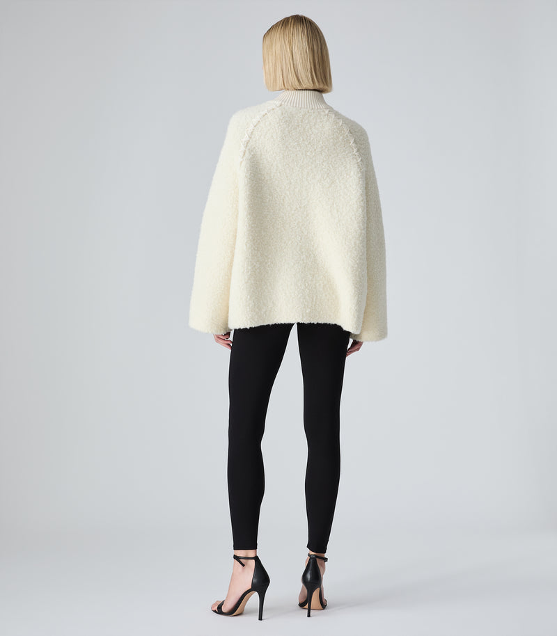 A model viewed from the back wearing a cream-colored, textured bouclé cape jacket, black leggings, and black strappy high heels against a light gray studio background.