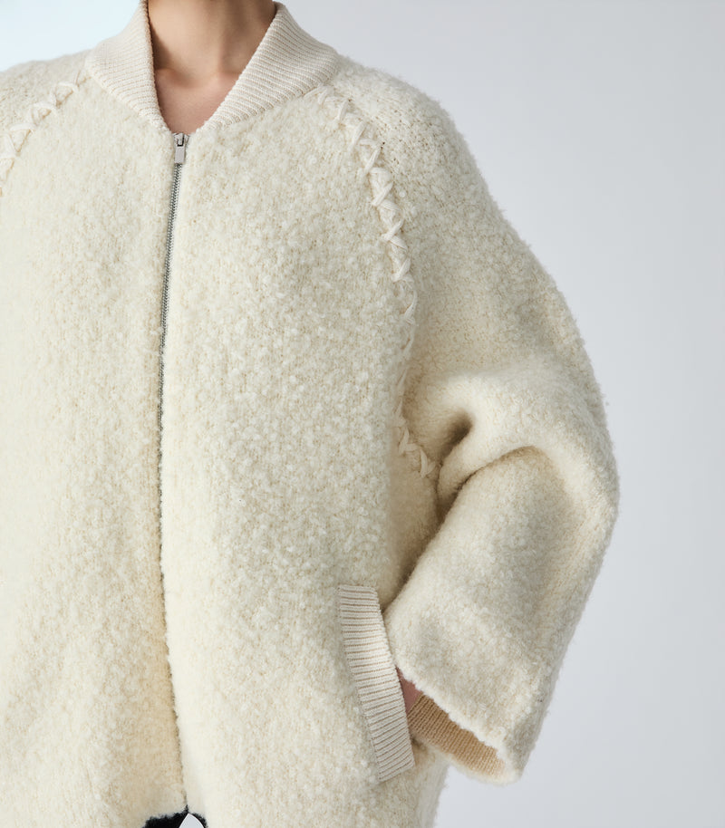 Close-up of a person wearing a cream-colored textured sherpa zip-up jacket with ribbed collar, cuffs, and hem, shown against a plain light gray background.