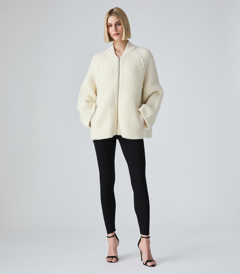 A woman modeling a cream-colored, textured teddy fleece zip-up jacket with black leggings and black high-heeled sandals, standing against a light gray studio background.