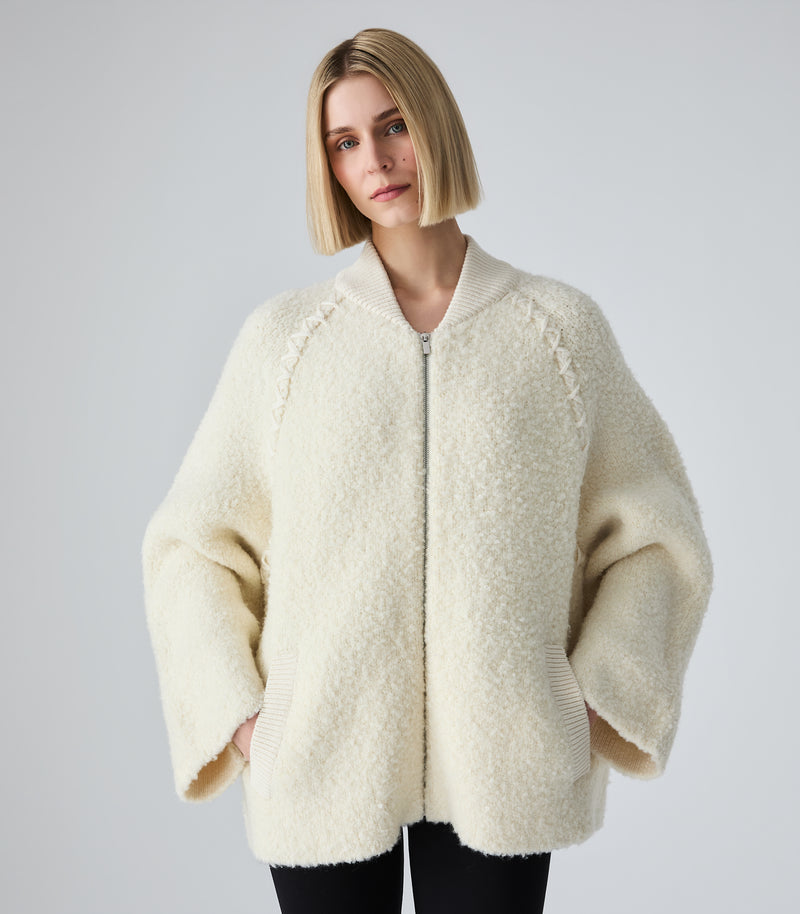 A woman with a short blonde bob wearing a cream-colored, textured teddy-bear zip-up jacket with wide balloon sleeves, set against a plain light gray studio background.