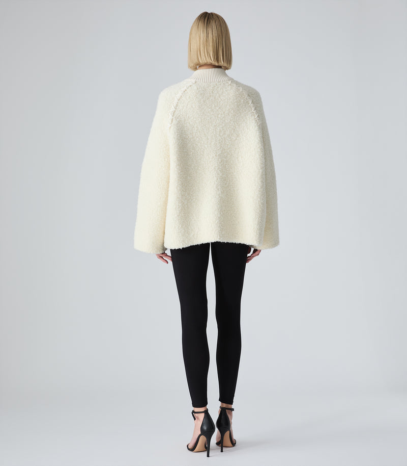 A woman viewed from behind wearing a cream-colored textured cropped jacket, black leggings, and black high-heeled sandals against a light gray studio background.