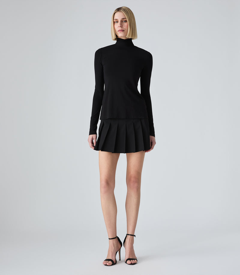 A woman modeling a black long-sleeve mock-neck mini dress with a pleated skirt, paired with black strappy high-heeled sandals, against a light gray studio background.