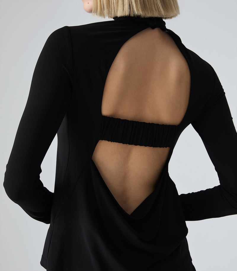 A woman shown from the back wearing a black long-sleeve top with a large open back design and a gathered elastic band across the upper back, against a light gray studio background.