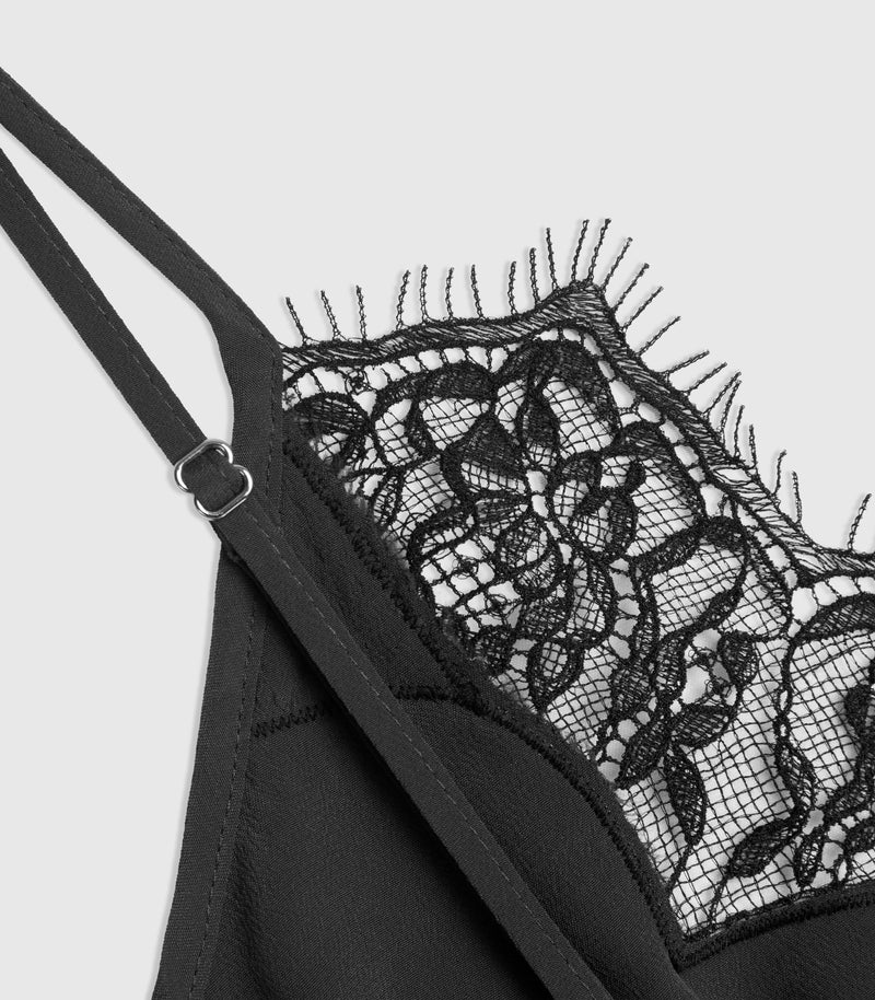Close-up of a black lace dress, showing the cup, thin shoulder straps with a silver buckle, and a detailed black floral lace trim with fringed edges against a light gray background.