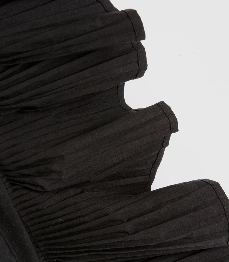 Close-up of a black pleated garment, showing the texture and layered ruffle construction against a plain light gray background.
