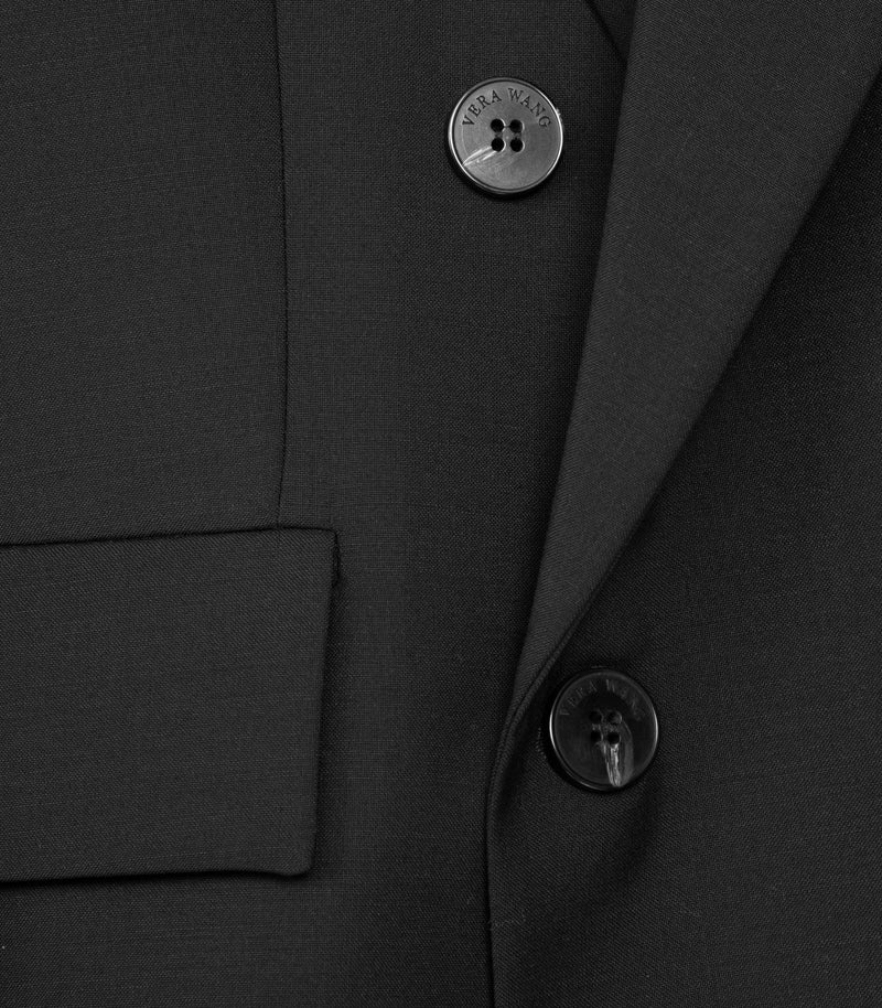 Macro close-up of the front fastening on a black double-breasted tailored blazer, highlighting the fine wool-blend fabric. The shot focuses on two stacked four-hole buttons engraved with the 'VERA WANG' logo, positioned alongside a sharp peak lapel edge and a horizontal lower flap pocket.