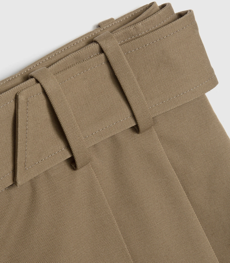 Close-up of a pair of pleated trousers in a solid taupe-brown fabric, showing the belt loops and waistband stitching against a light gray background.
