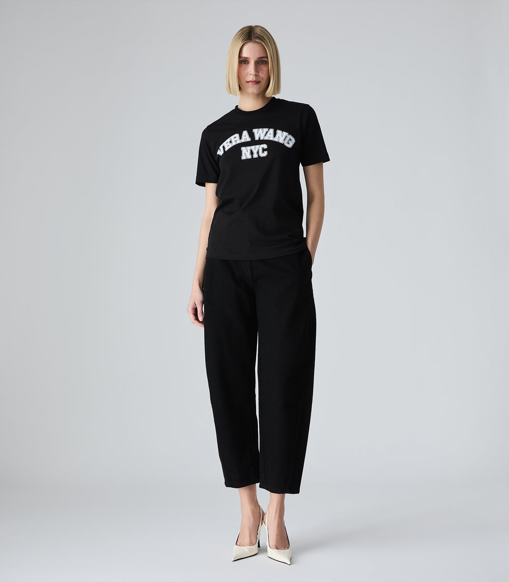 Female model wearing a slim-fit black t-shirt with 'VERA WANG NYC' spell-out logo across the front, paired with black trousers and white heels, against a white background.