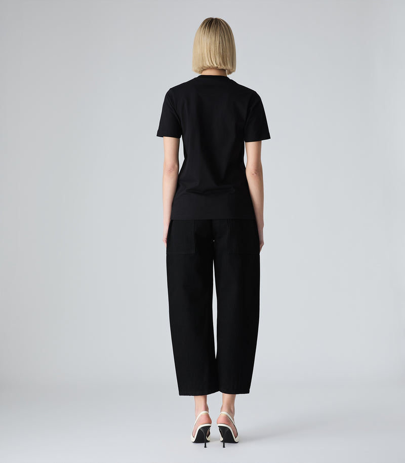 Female model wearing a slim-fit black t-shirt paired with black trousers and white heels, against a white background.