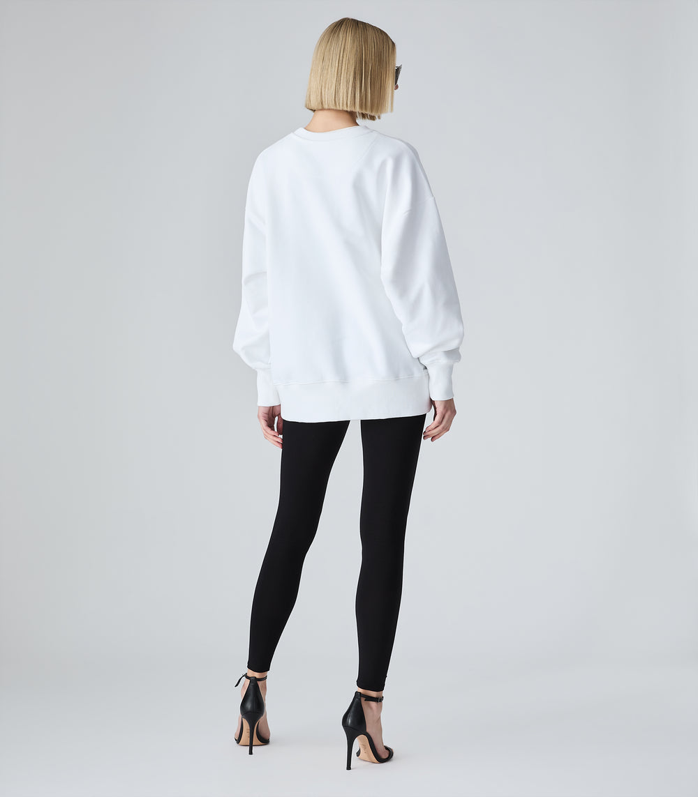 Female model wearing a white crewneck sweatshirt, paired with black leggings and heels, against a white background.