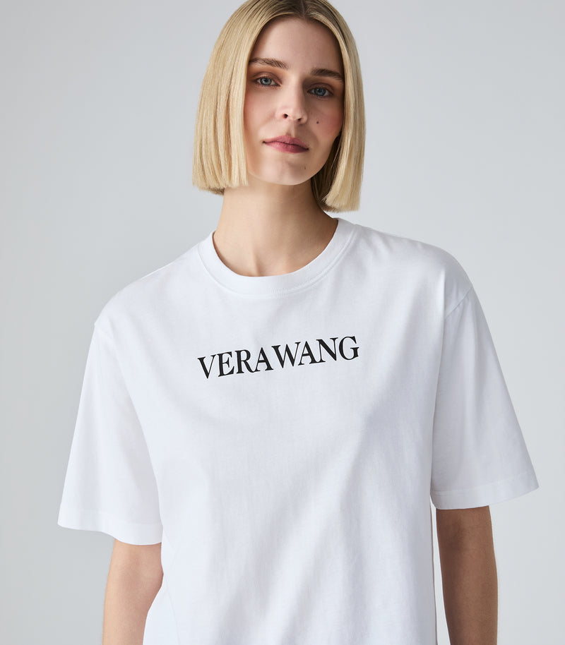 Half-length view of a model wearing an oversized, loose-fit white t-shirt featuring the classic 'VERA WANG' brand logo printed in black across the upper chest, designed with wide short sleeves and a crew neckline, against a light grey background.