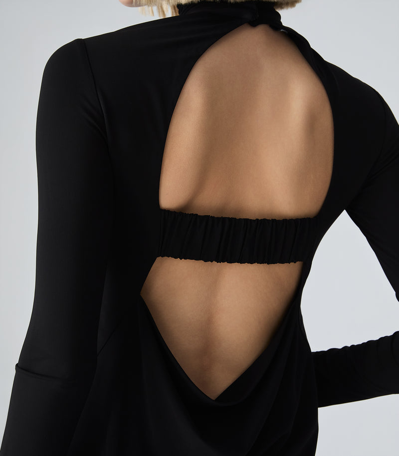 Close-up back view of a model wearing a black long-sleeve top with a large open back and a gathered elastic band across the upper back, against a light gray studio background.