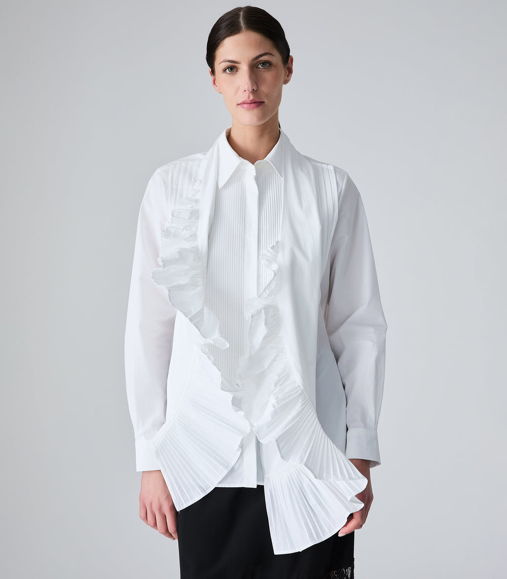 A woman modeling a white long-sleeve blouse with pleated detailing, paired with a white pleated scarf with a ruffle trim and black pants, against a light gray studio background.