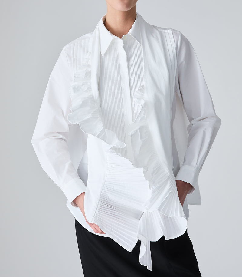 A woman modeling a white long-sleeve blouse with pleated detailing, paired with a white pleated scarf with a ruffle trim and black pants, against a light gray studio background.