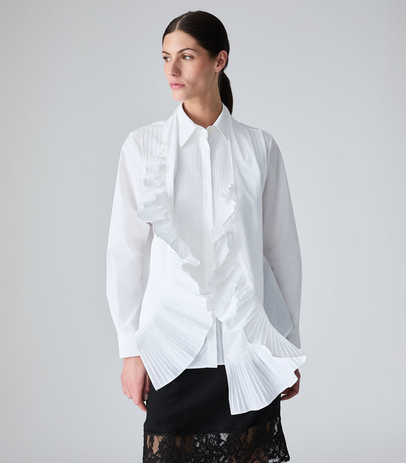 A woman modeling a white long-sleeve blouse with dramatic pleated and ruffled scarf, paired with a black skirt, against a plain light gray studio background.
