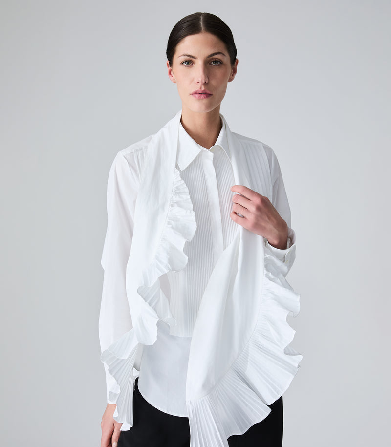 A woman modeling a white long-sleeve blouse with pleated details, paired with a white scarf that has dramatic ruffle and pleated details, worn by a model against a light gray studio background.