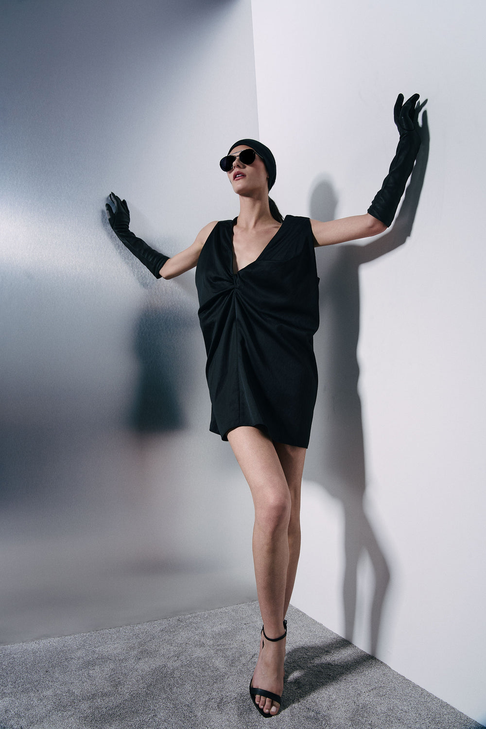 A woman posing in a black wrap-style mini dress, long black gloves, and black sunglasses, leaning against a white wall in a studio setting.