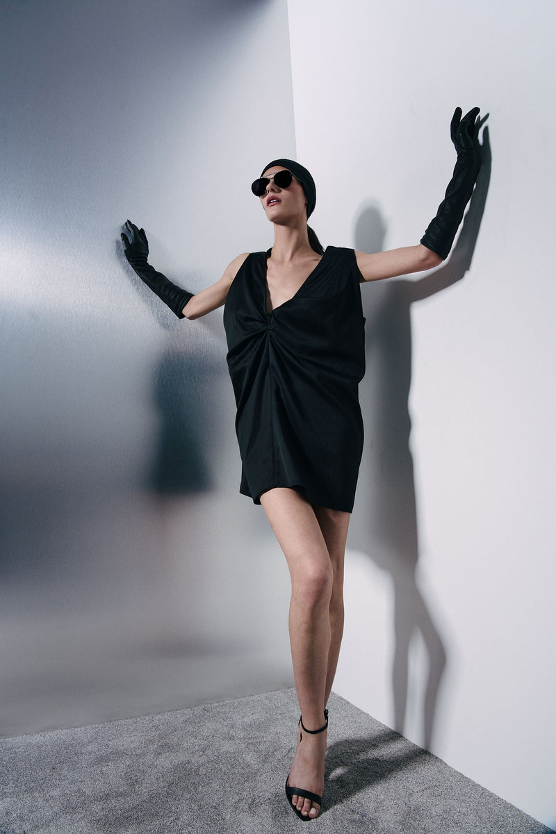A woman posing in a black wrap-style mini dress, long black gloves, and black sunglasses, leaning against a white wall in a studio setting.