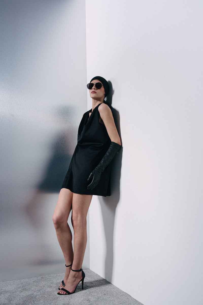 A model wearing a black sleeveless mini dress, black long gloves, black sunglasses, and black high-heeled sandals leans against a white wall in a minimalist interior setting.