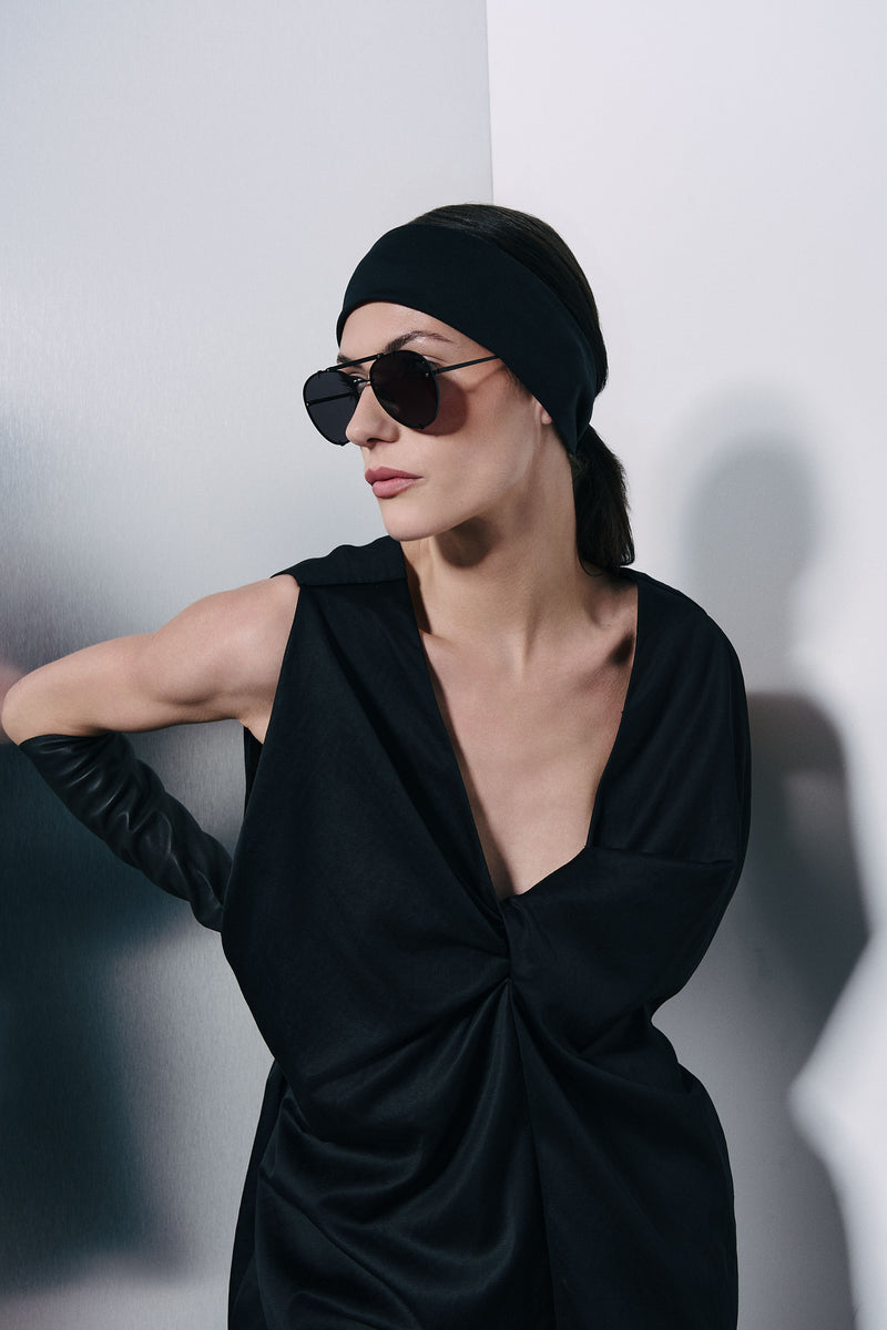 A woman wearing a Winona Black Short Dress from Vera Wang, with a twisted front detail, paired with a black headband, and dark sunglasses, posed against a light gray wall with soft shadows.