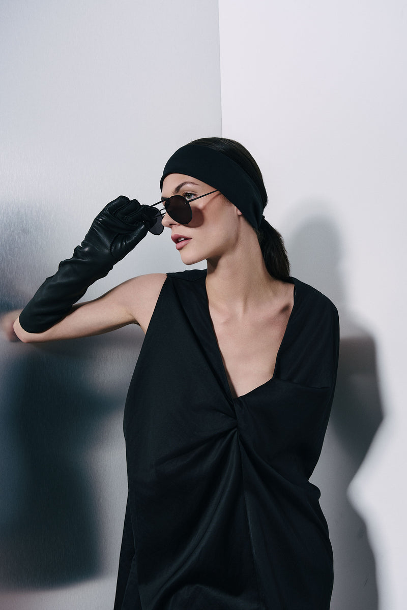 A woman wearing a black draped wrap dress, a black headband, black gloves, and dark sunglasses, posing against a minimalist white wall with soft shadows.